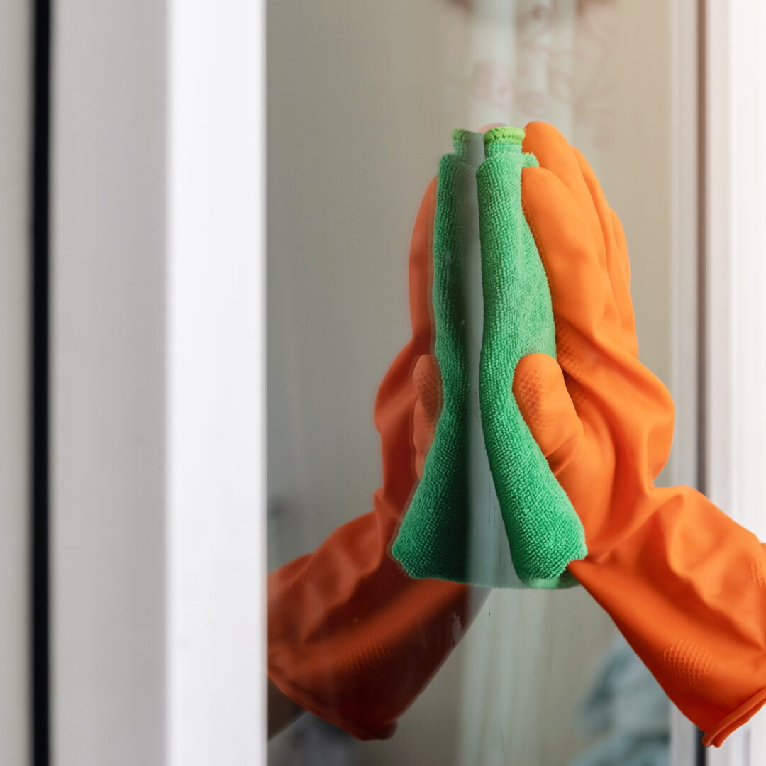 Women's hand wears orange rubber gloves, using green cloth to wipe the glass of the building with a glass cleaner.