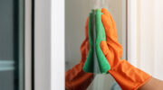 Women's hand wears orange rubber gloves, using green cloth to wipe the glass of the building with a glass cleaner.