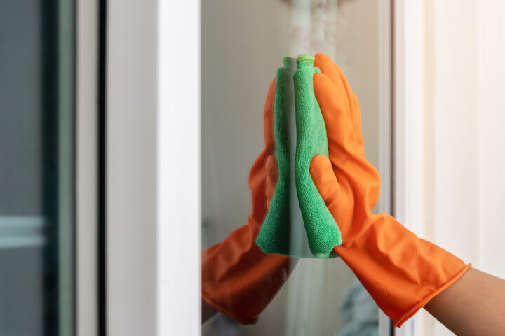 Women's hand wears orange rubber gloves, using green cloth to wipe the glass of the building with a glass cleaner.