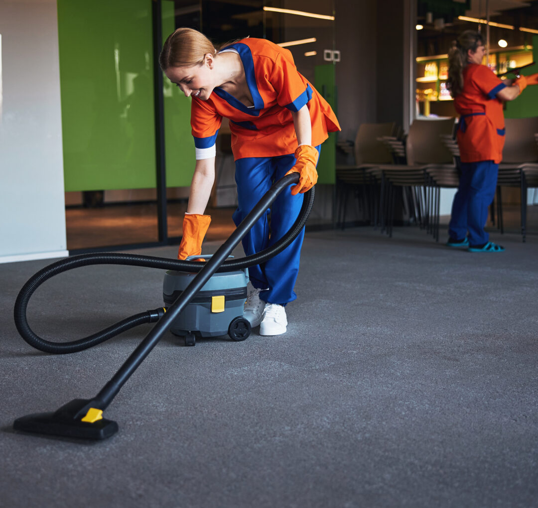 Smiling young female worker in the uniform leaning over a corded bagless canister vacuum cleaner