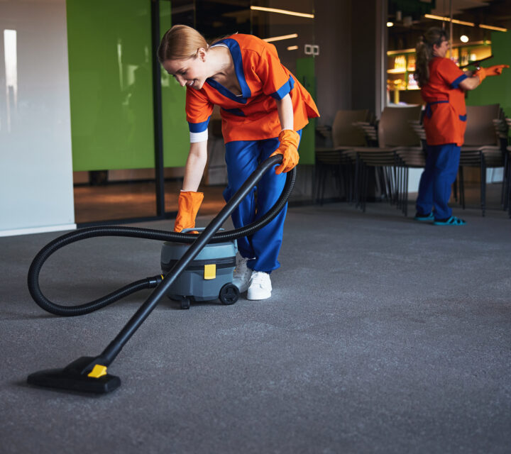 Smiling young female worker in the uniform leaning over a corded bagless canister vacuum cleaner