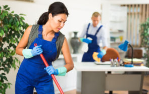 Portrait of professional female worker of cleaning service wearing uniform and rubber gloves wiping office floor with mop