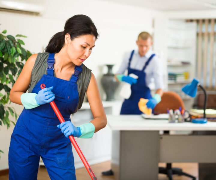 Portrait of professional female worker of cleaning service wearing uniform and rubber gloves wiping office floor with mop