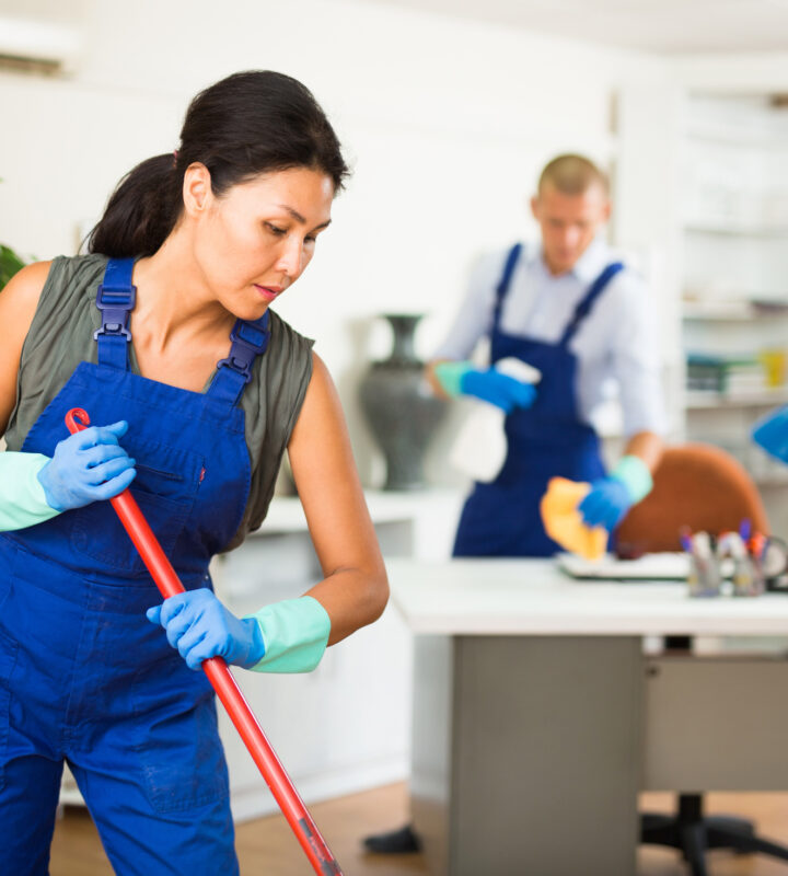 Portrait of professional female worker of cleaning service wearing uniform and rubber gloves wiping office floor with mop